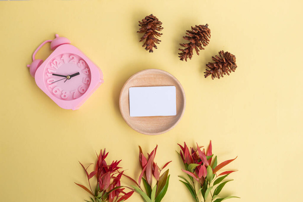 Tarjeta de visita sobre madera con flores de pino y reloj despertador sobre fondo amarillo pastel. estilo minimalista - Foto, Imagen