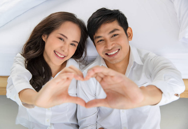 Młoda, atrakcyjna azjatycka para w czystej białej koszuli, siedząca razem na podłodze w sypialni i patrząca w górę na kamerę, robiąca znak serca. Pojęcie miłości i szczęśliwego związku. - Zdjęcie, obraz