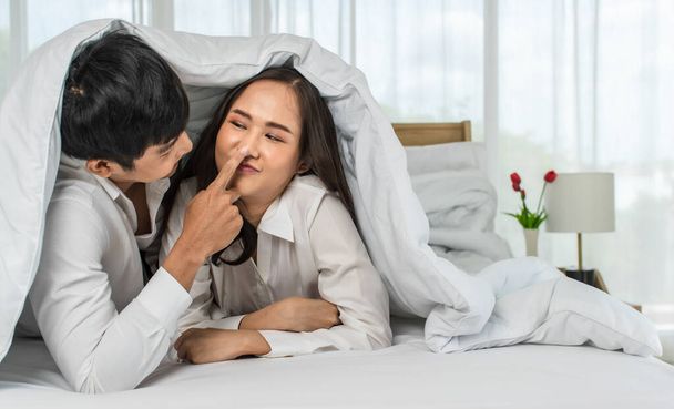 若い幸せな魅力的なアジアのカップルは、白いカーテンの背景を持つベッドでお互いにからかっている。愛と幸せな関係の概念. - 写真・画像