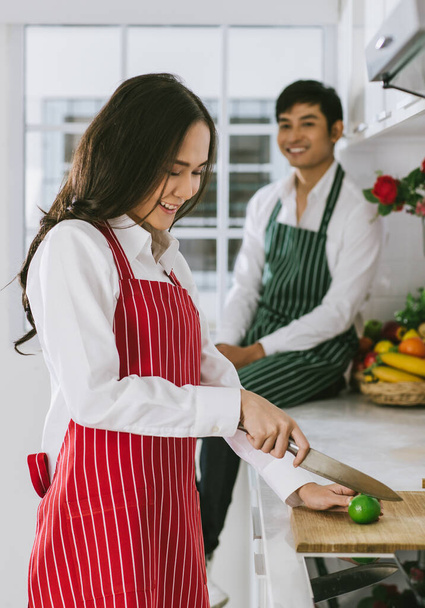 Junge attraktive asiatische Paare in der Küche. Frau in roter Schürze mit Zitronenscheiben bereitet sich auf das Kochen vor und Mann sitzt im Hintergrund in grüner Schürze. Konzept für gesunde und glückliche Liebe und Kochen. - Foto, Bild