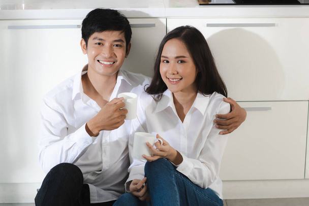 Młoda atrakcyjna azjatycka para w białej koszuli i dżinsach siedząca razem w białej kuchni trzymająca białe kubki kawy i uśmiechająca się do siebie. Koncepcja zdrowej i szczęśliwej miłości i gotowania. - Zdjęcie, obraz