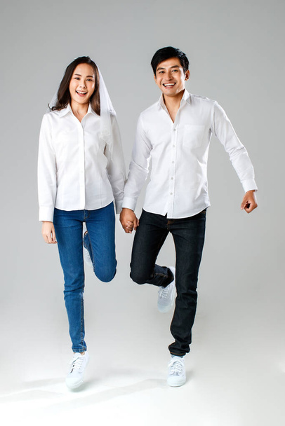Młoda, atrakcyjna azjatycka para w białej koszuli i welonie, trzymająca się za ręce, skacząca i uśmiechnięta na białym tle. Koncepcja fotografii przedślubnej. Izolacja. - Zdjęcie, obraz