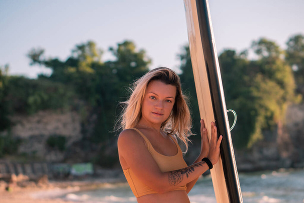 Portrait of young blonde surfer girl with long surf board single fin on the beach at golden hour, sunset in Bali - Foto, Bild