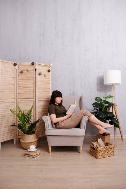 Woman reading a book in armchair with copy space on concrete wall - Photo, image