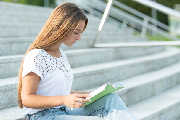 Hübsche Studentin mit langen blonden Haaren sitzt auf der Treppe und liest ein Buch. - Foto, Bild