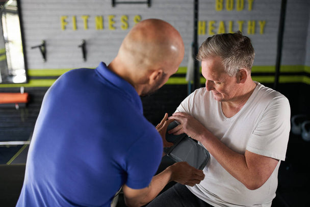 男性の健康と健康パーソナルトレーナーは、家庭用ジムの小さなビジネスでのトレーニング前に腕の上に血圧モニタをフィッティングで成熟したシニア男性を支援 - 写真・画像