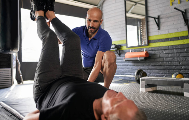 Male personal trainer assisting an elderly mature man in doing raised leg ab crunches on mat. Working on core strength and balance. Working out at home gym. Small business - Photo, Image