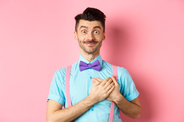 Χαρούμενος νεαρός άνδρας με μουστάκι, φοράει πουκάμισο και παπιγιόν, κρατά τα χέρια στην καρδιά και χαμογελά ευγνώμων, λέγοντας ευχαριστώ, στέκεται σε ροζ φόντο - Φωτογραφία, εικόνα