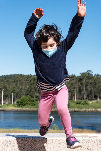 caucasian child jump at grass from the bench on the promenade with smile face behind the mask. - Photo, image