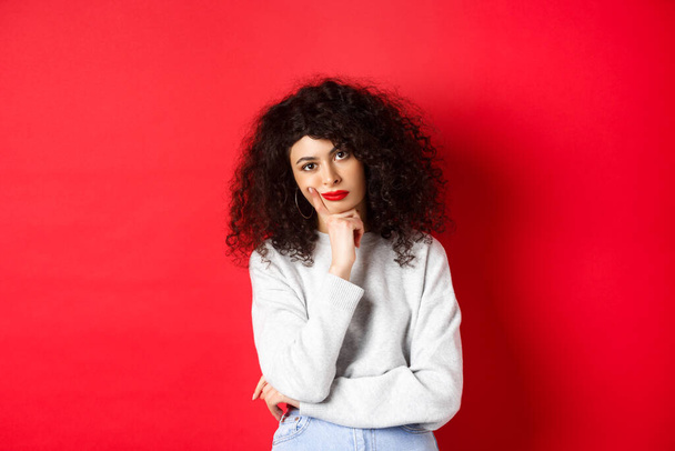 Bored and tired curly woman with red lips, looking bothered at camera, standing against studio background - Photo, Image