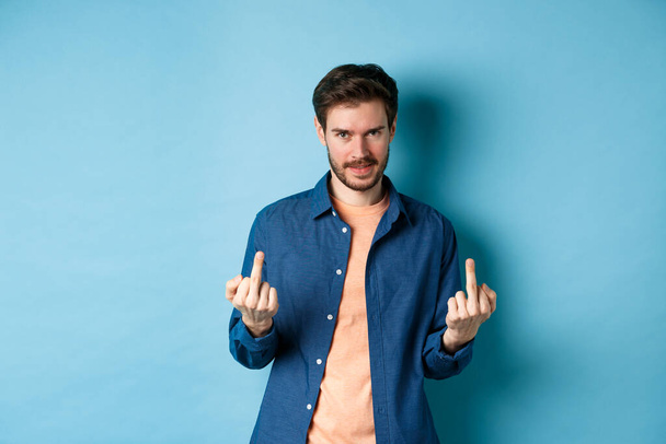 Ignorant and unbothered man showing middle-fingers and looking reluctant at camera, give zero fucks, standing on blue background - Foto, immagini