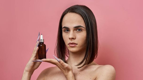Beautiful young transgender with dark brown hairstyle looking at camera, holding bottle of hyaluronic acid or oil for skincare, posing isolated over pink background. Beauty, lgbtq, generation Z - 写真・画像