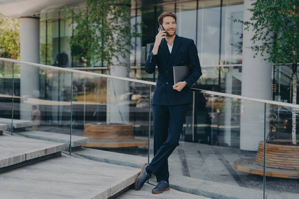 Full length shot of elegant businessman in formal clothes has telephone conversation holds digital gadget under arm answers phone call stands outdoor near office building or business center. - Foto, Imagen
