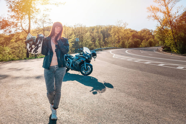 Young smiling woman in a leather jacket, holding a helmet in her hand. In the background, a motorcycle and a road. Motorcycle local travel concept. - Fotografie, Obrázek