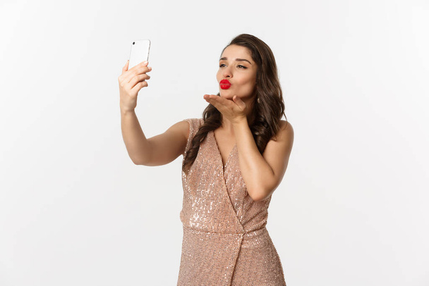 Красивая женщина с макияжем и прической, одетая в гламурное платье, делающая селфи на смартфоне, празднующая новогоднюю вечеринку, белый фон - Фото, изображение