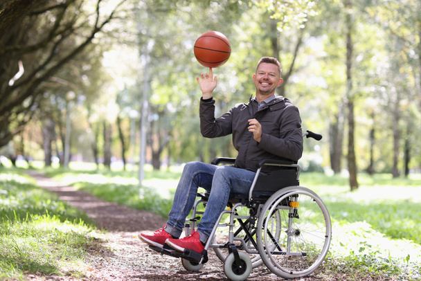 Sonriendo hombre discapacitado gira pelota de baloncesto en su dedo mientras está sentado en silla de ruedas en el parque. Concepto de deporte para personas con discapacidad - Foto, Imagen