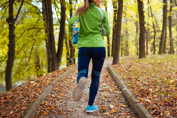 Runner working out in autumn park wearing sportive clothes. Young woman running with water bottle among yellow trees. Close up of shoes - Foto, Imagem