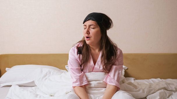Dormir mujer de pelo oscuro en elegante pijama rosa con máscara especial negro ojo dormido, se levanta rápidamente se sienta en la cama expresando su insatisfacción contra la pared blanca en el primer plano del dormitorio - Foto, Imagen