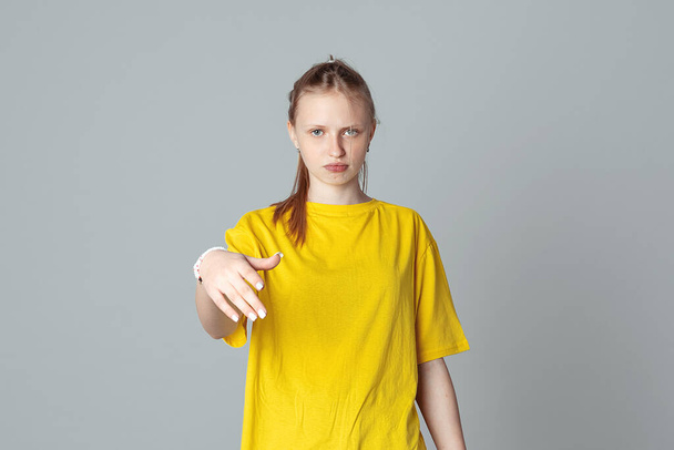 Portrait of displeased teen girl gestures by hand isolated on light grey background, wearing in casual blank yellow t shirt. Stop, no, stay back - Photo, Image