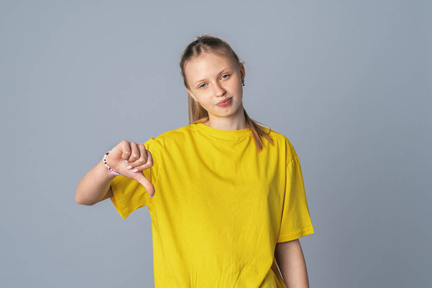 Beautiful teen girl feeling displeased, showing thumb down, isolated on light gray background, dressed in casual yellow t shirt. Expressive facial expressions - Photo, Image
