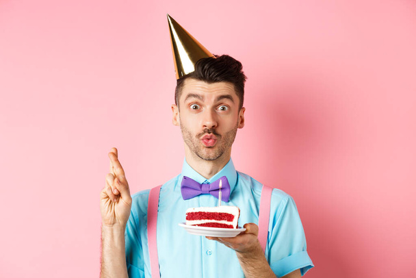 Vacaciones y concepto de celebración. Hombre lindo y divertido haciendo deseo de cumpleaños con los dedos cruzados, sosteniendo el pastel del día b y mirando soñador a la cámara, fondo rosa. - Foto, imagen