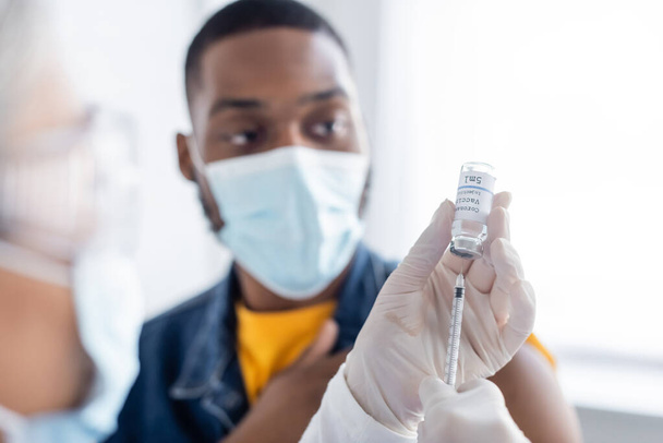 verpleegkundige in latex handschoenen vullen spuit met coronavirus vaccin in de buurt van wazig Afro-Amerikaanse man met medisch masker - Foto, afbeelding