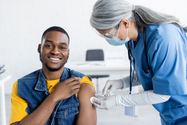 Ασιάτης γιατρός με ιατρική μάσκα που εμβολιάζει έναν ευτυχισμένο Αφροαμερικανό στην κλινική. - Φωτογραφία, εικόνα