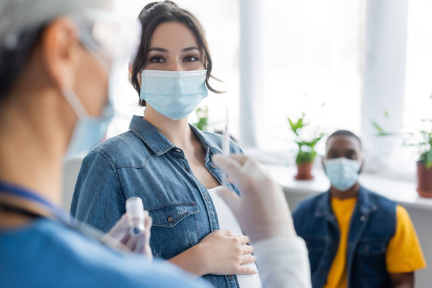verschwommene Krankenschwester hält Spritze und Gefäß mit Impfstoff in der Nähe von schwangerer Frau und afrikanisch-amerikanischem Mann in medizinischen Masken - Foto, Bild