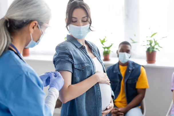 ώριμη νοσοκόμα εμβολιάζοντας έγκυο γυναίκα κοντά θολή αφροαμερικανός άνδρας με ιατρική μάσκα - Φωτογραφία, εικόνα