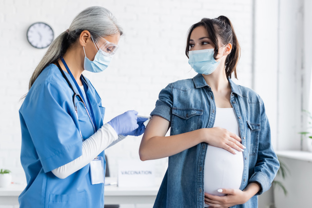 mujer asiática de mediana edad en máscara médica y guantes de látex vacunando a la mujer embarazada - Foto, Imagen