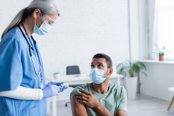 Dojrzała azjatycka pielęgniarka w masce medycznej, goglach i rękawiczkach lateksowych ze szczepionką w pobliżu młodego mężczyzny - Zdjęcie, obraz