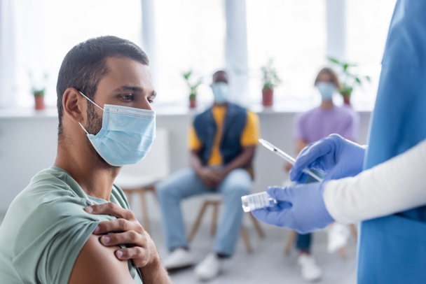 enfermeira em luvas de látex segurando seringa e frasco com vacina perto do homem em máscara médica e pessoas multiétnicas borradas - Foto, Imagem