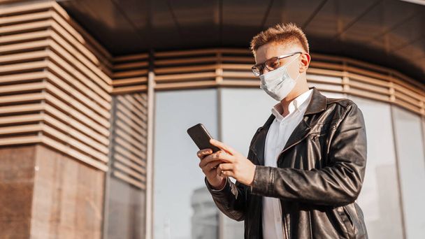 Handsome man in a protective mask on his face with glasses with a smartphone on the street of a big city. Businessman talking on the phone on urban background - Photo, image