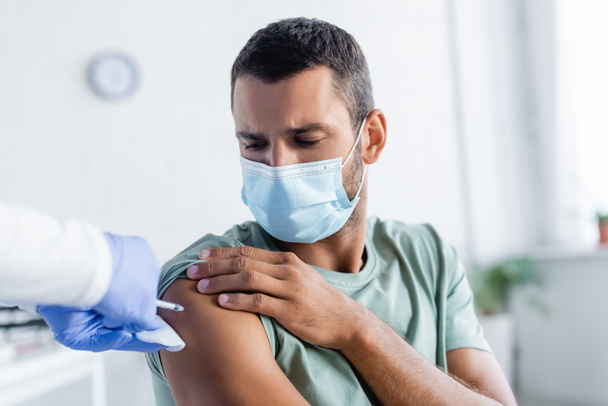 νοσοκόμα με γάντια λάτεξ που εμβολιάζει νεαρό άνδρα με ιατρική μάσκα - Φωτογραφία, εικόνα