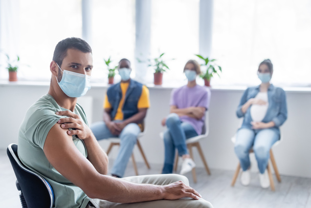 молодий чоловік у захисній масці дивиться на камеру біля розмитих багатоетнічних людей, що сидять у клініці, концепція вакцинації
 - Фото, зображення