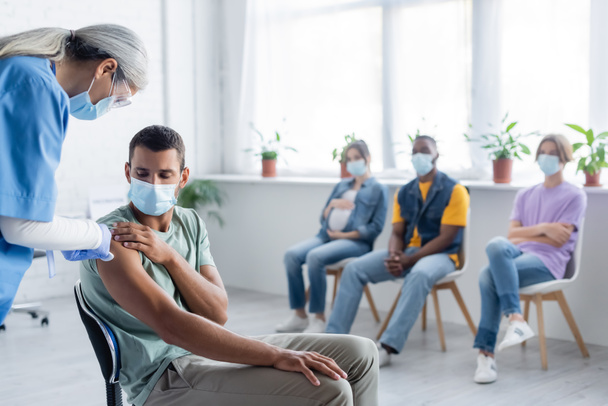mediana edad asiático enfermera dando vacuna inyección a joven hombre y pacientes en médico máscaras - Foto, Imagen