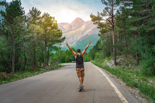 Hipster guy with long board with open arms enjoying life in the middle of a mountain road and a beautiful landscape. - Foto, imagen