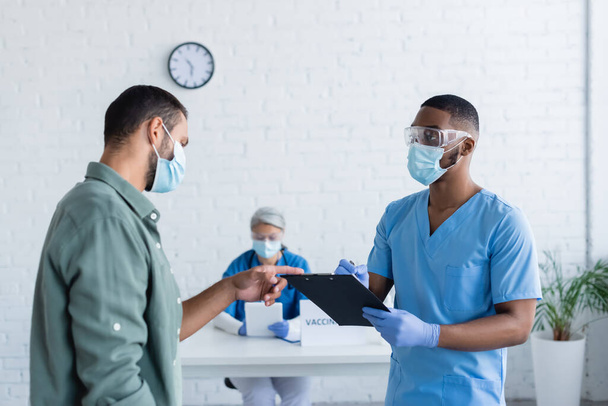 man in medical mask pointing at clipboard in hands of african american doctor in vaccination center - 写真・画像