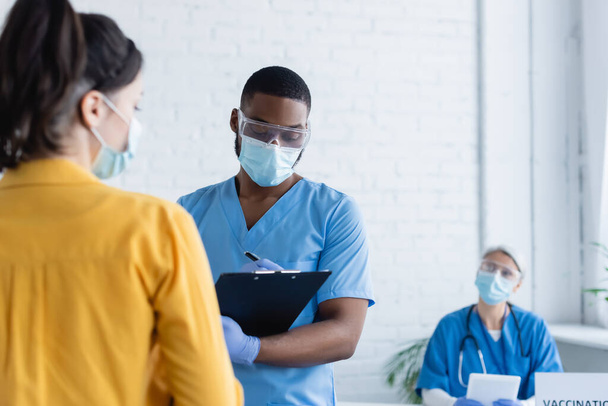 african american man in medical mask writing on clipboard near blurred woman in vaccination center - Foto, Imagen