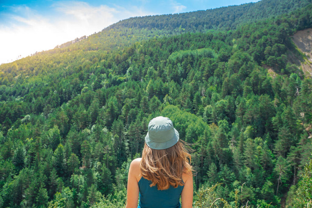 Young woman standing alone outdoors with wild forest mountains in background. Traveling lifestyle and survival concept rear view. Trekking with a green hat. - Foto, imagen
