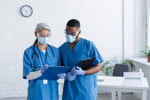 mature doctor in medical mask writing on clipboard near african american colleague, vaccination concept - Foto, Bild