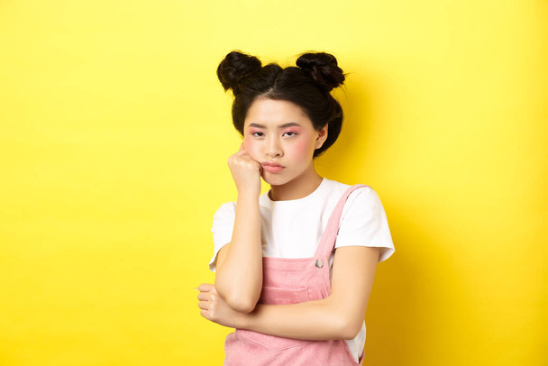 Bored asian teen girl with stylish makeup and summer clothes, looking reluctant and indifferent, standing on yellow background. - Photo, Image