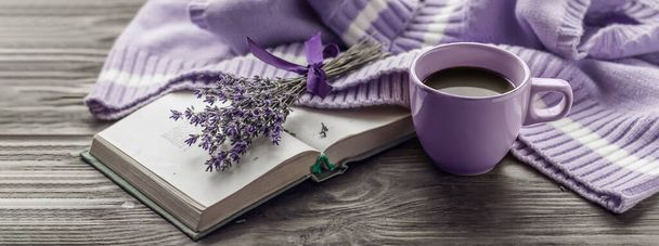 Café matutino. Una taza de café sobre una mesa de madera, un libro abierto y un suéter caliente sobre el fondo de un ramo de flores. Bodegón en color lavanda. Acogedora mañana. - Foto, imagen