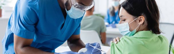 病院で若い女性を予防接種する医療マスクのアフリカ系アメリカ人医師バナー - 写真・画像