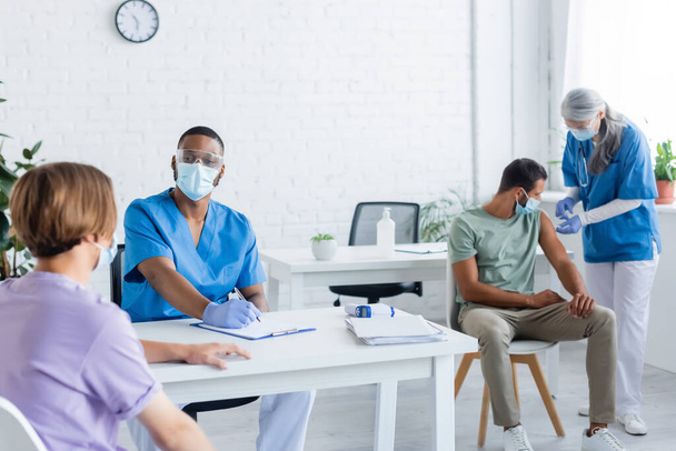 interracial doctors in medical masks and uniform working with young men in vaccination center - Photo, Image