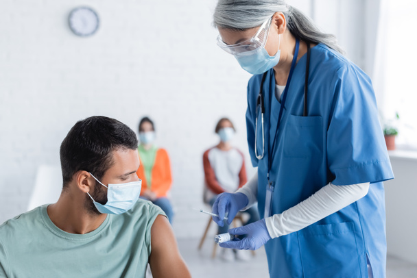 volwassen aziatische arts in medisch masker met spuit en pot met vaccin in de buurt van jonge man en wazig patiënten - Foto, afbeelding
