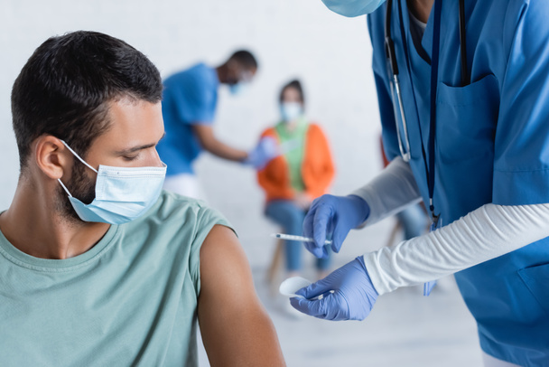 médico en guantes de látex que sostiene la jeringa cerca del hombre en máscara médica, concepto de vacunación - Foto, Imagen
