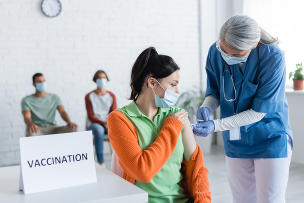 dojrzewająca pielęgniarka wstrzykująca młodej kobiecie w pobliżu liter szczepień i niewyraźnych pacjentów w maskach medycznych - Zdjęcie, obraz