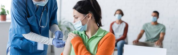медсестра в латексних рукавичках, що вакцинує молоду жінку в медичній масці біля розмитих пацієнтів, банер
 - Фото, зображення