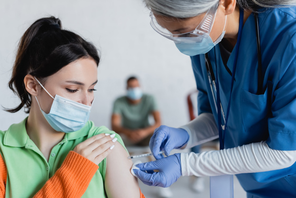 dojrzała azjatycka pielęgniarka w masce medycznej, goglach i rękawiczkach lateksowych szczepiących młodą kobietę - Zdjęcie, obraz